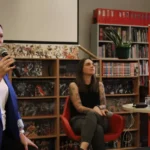Kobieta w niebieskiej marynarce przemawia do mikrofonu, a dwie inne kobiety siedzą obok niej podczas dyskusji panelowej w bibliotece.