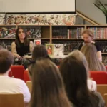 Dwie kobiety siedzą przy stole przed regałem z książkami i przemawiają przez mikrofony do publiczności zgromadzonej w sali wykładowej.