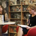 Dwie kobiety siedzą na czerwonych krzesłach przy stole, na którym leżą książki; jedna z nich mówi do mikrofonu, trzymając w ręku otwarty notatnik.