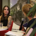 Kobieta z wytatuowanymi ramionami siedzi przy stole i słucha, jak inna osoba przemawia do mikrofonu podczas wydarzenia organizowanego w bibliotece.