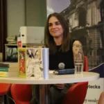 Kobieta siedząca przy stole z książkami, przemawiająca do mikrofonu podczas wydarzenia.