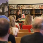 Kobieta przemawia do mikrofonu, siedząc przy biurku przed regałem bibliotecznym, twarzą zwróconą w stronę publiczności zgromadzonej w sali wykładowej.