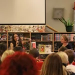 Kobieta z tatuażami przemawia do mikrofonu, siedząc przy stole wraz z inną kobietą przed publicznością zgromadzoną w bibliotece.