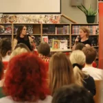 Dwie kobiety przemawiają do mikrofonów podczas wydarzenia literackiego odbywającego się przed regałem z książkami, a publiczność obserwuje je z miejsc.