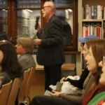 Mężczyzna w garniturze przemawia do mikrofonu przed publicznością siedzącą w bibliotece.