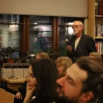 Mężczyzna w marynarce stoi i przemawia do mikrofonu przed zgromadzoną w bibliotece publicznością.