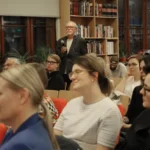 Mężczyzna przemawiający do mikrofonu stoi przed publicznością siedzącą w bibliotece.