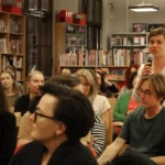Kobieta w białej bluzce przemawia do ręcznego mikrofonu, stojąc przed publicznością w bibliotece.