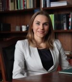 Osoba z długimi włosami siedzi w biurze ubrana w jasną marynarkę i ciemny top. Uśmiecha się i siedzi przy biurku, a w tle widać regały z książkami.