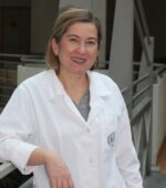 Osoba ubrana w biały fartuch laboratoryjny stoi na korytarzu, opierając się o poręcz. Ma krótkie włosy i uśmiecha się do kamery. Wnętrze ma jasne ściany i poręcze.