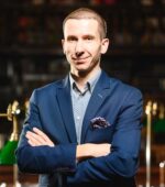 Osoba z krótkimi włosami ubrana w niebieski garnitur i wzorzystą koszulę pewnie stoi ze skrzyżowanymi ramionami. Tło jest ciemne i zawiera rozmyte regały na książki i zielone lampy.