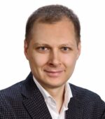 Mężczyzna w teksturowanej marynarce i białej koszuli uśmiecha się do kamery na białym tle.