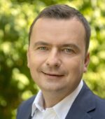 Mężczyzna w garniturze i białej koszuli uśmiecha się do kamery. Tło jest rozmyte, zielone, liściaste, sugerując środowisko zewnętrzne.