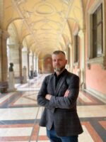 Mężczyzna z brodą pewnie stoi ze skrzyżowanymi ramionami w długim, łukowatym korytarzu. Sufit jest bogato zdobiony, a podłoga ma geometryczny wzór. Okna i posągi zdobią korytarz.