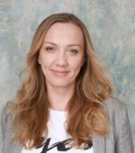 Kobieta o długich blond włosach uśmiecha się łagodnie, ubrana w białą koszulę z częściowo widocznym wzorem i jasnoszary blezer. Tło to teksturowana mieszanka stonowanych odcieni niebieskiego i zieleni.
