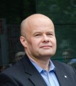 Łysy mężczyzna w niebieskiej koszuli i czarnym blezerze stoi na zewnątrz. Jego wyraz twarzy jest neutralny, patrzy lekko na bok. Tło stanowi odblaskowa powierzchnia szklana z widocznymi rozmazanymi postaciami.