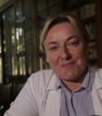 Osoba z krótkimi blond włosami się uśmiecha, ma na sobie biały płaszcz na koszuli z kołnierzykiem. Są w środku, z regałami na książki i szklanymi oknami w tle. Oświetlenie jest ciepłe i nastrojowe.