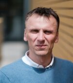 Mężczyzna z krótkimi włosami i niebieskim swetrem stoi na zewnątrz na tle ceglanego muru.