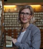 Kobieta w okularach i z blond włosami stoi przed ścianą kwadratowych drewnianych szuflad, trzymając otwarty katalog kartkowy. Ma na sobie szary blezer na białej koszuli i delikatnie uśmiecha się do kamery.